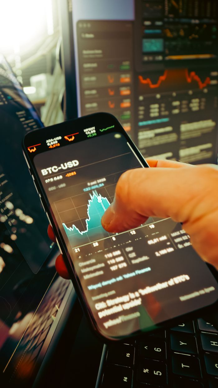 creative-01 A hand holds a smartphone displaying BTC-USD chart amidst digital trading screens, highlighting crypto finance.